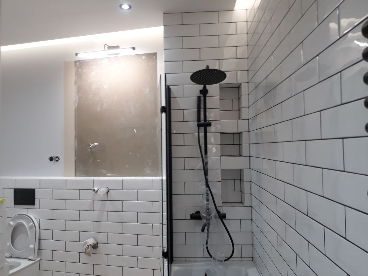 Biała łazienka w trakcie wykańczania, widoczne białe płytki ułożone w cegiełkę, czarna deszczownica w folii ochronnej, przygotowane miejsce pod lustro, częściowo zamontowana toaleta.