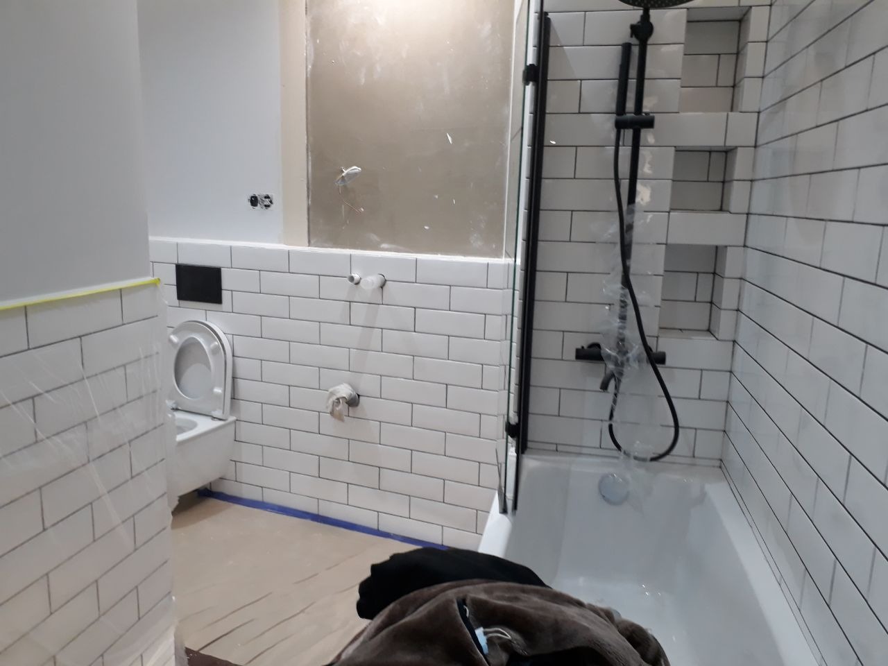 Łazienka w trakcie remontu: białe, prostokątne płytki na ścianach, częściowo zabezpieczone folią, czarna armatura prysznicowa, wisząca toaleta, widoczne przygotowania do malowania ściany.