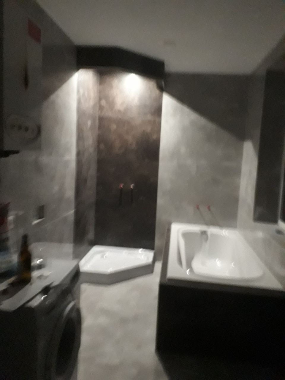 Nowoczesna łazienka w szarościach z kabiną prysznicową i obudowaną wanną, oświetlenie punktowe, widoczna pralka i butelka na blacie.