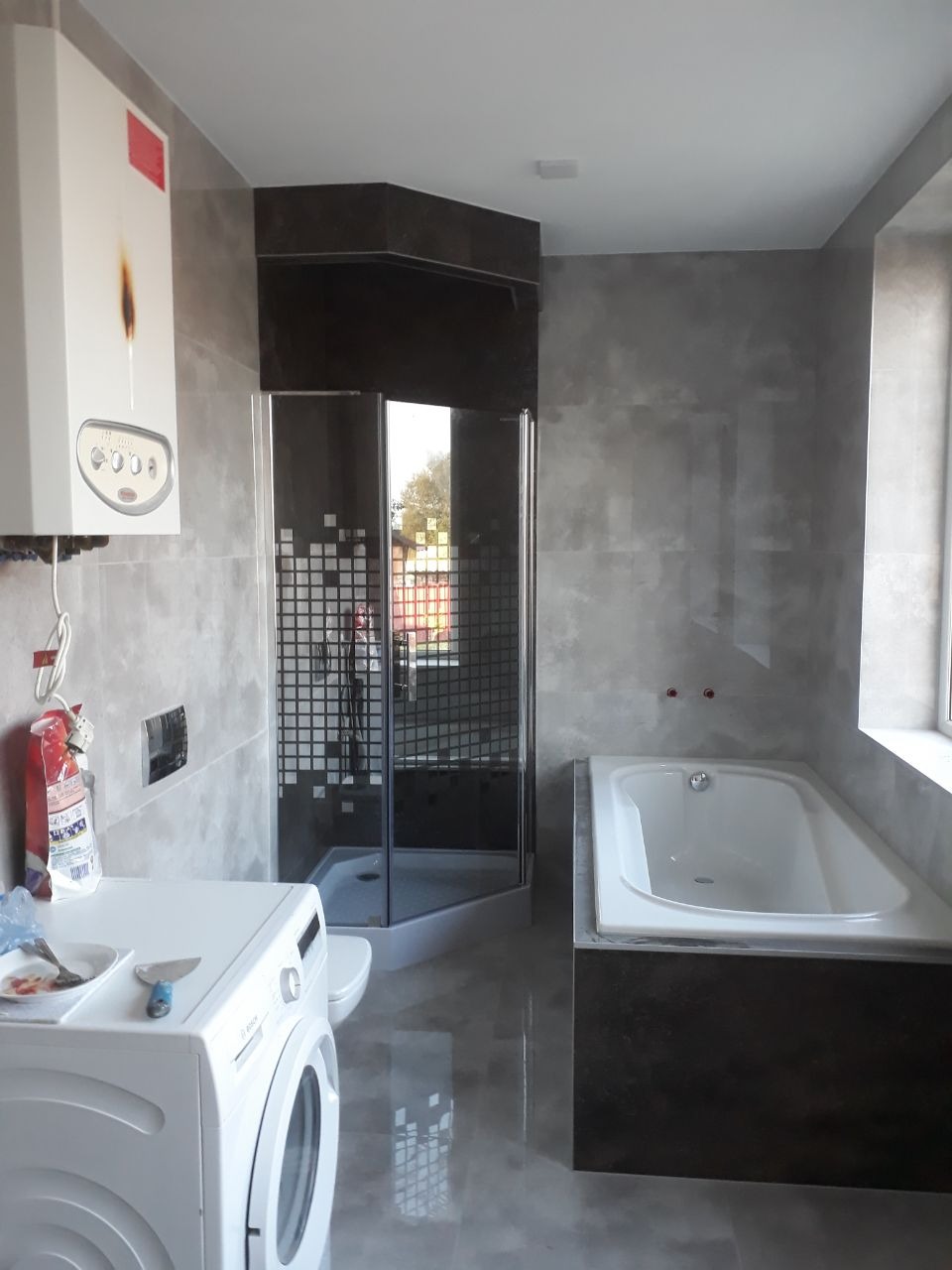 Wykończona łazienka z szarą płytką imitującą beton, narożną kabiną prysznicową z czarnymi akcentami i białą wanną obudowaną ciemnym gresem, widoczna pralka i piec gazowy, na pralce narzędzia...