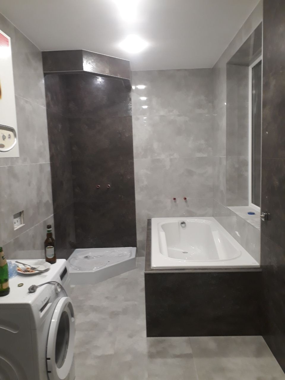 Wykończona łazienka z szaro-brązowymi płytkami imitującymi kamień, białą wanną i brodzikiem bez kabiny, pralka z niedojedzonym posiłkiem na wierzchu, butelki po piwie.