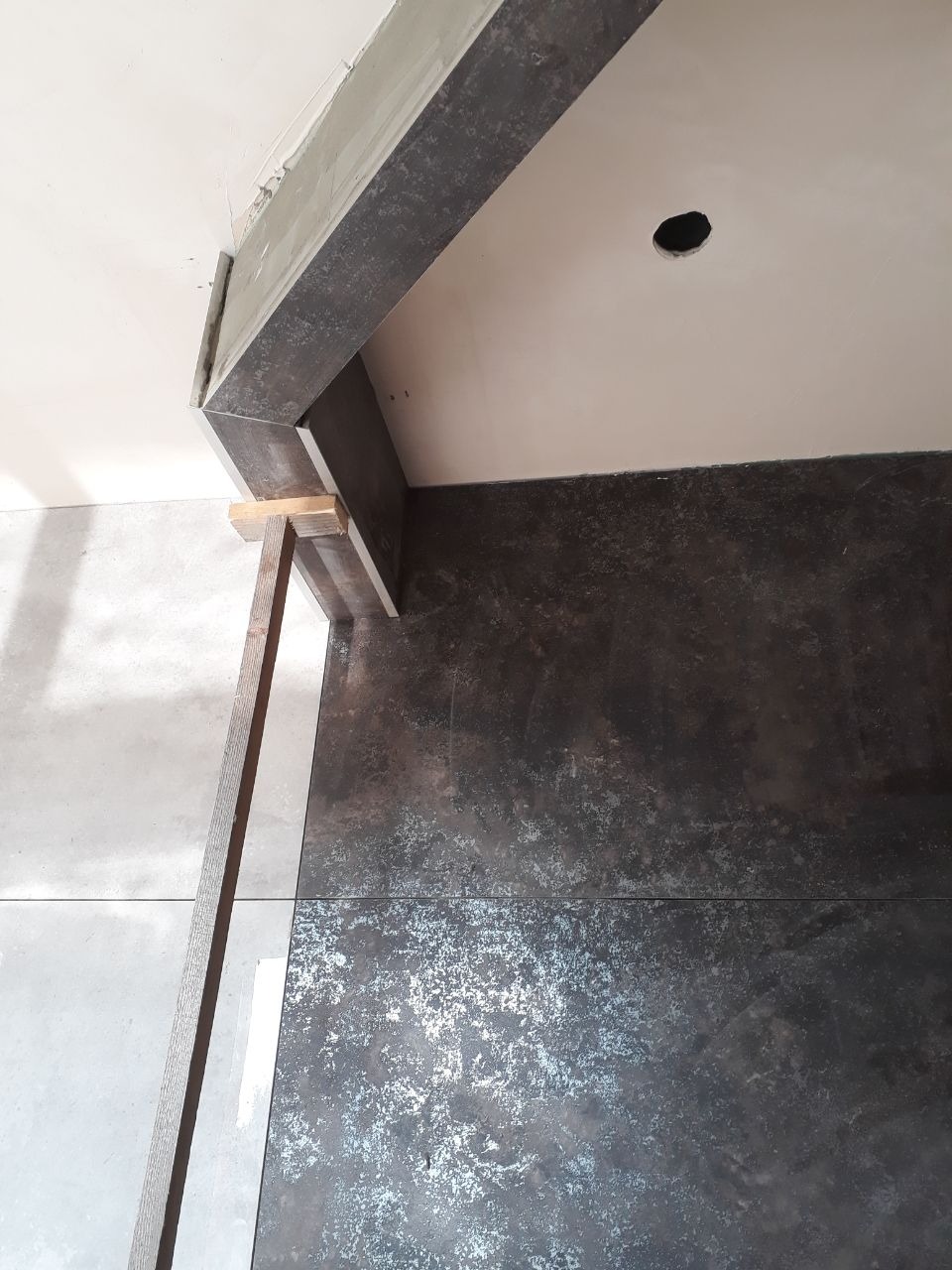 Ciemne płytki podłogowe o metalicznym połysku, ułożone w duży format, kontrastujące z jasnymi płytkami na ścianie, drewniana listwa przypodłogowa w trakcie montażu, widoczny fragment sufitu...