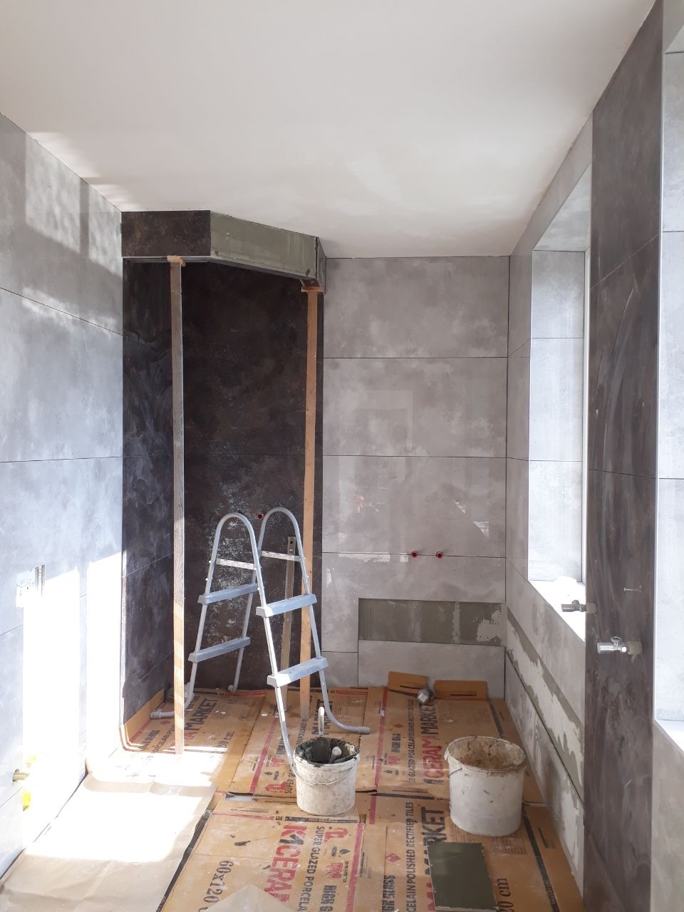Łazienka w trakcie remontu: szare płytki na ścianach, drabina oparta o ścianę obłożoną ciemnym materiałem, dwa wiadra z zaprawą na podłodze zabezpieczonej kartonem, widoczne instalacje wodne.