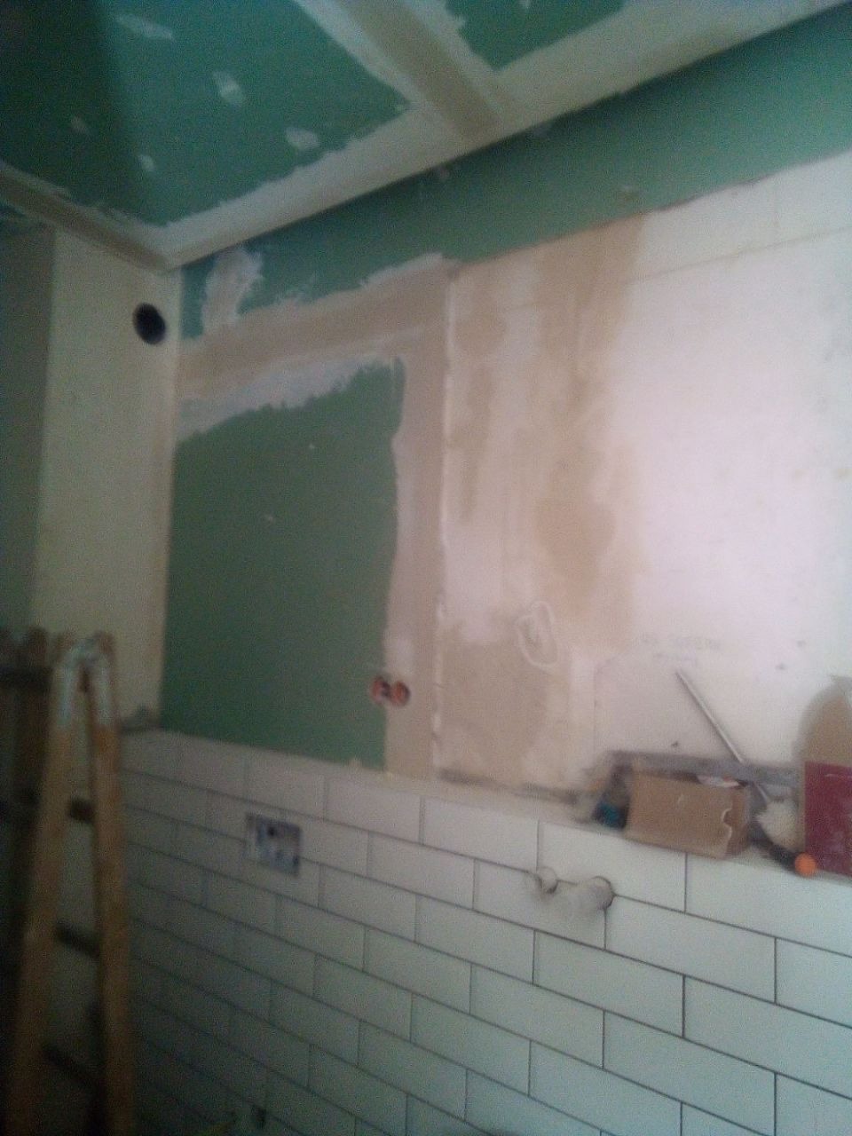 Ściana w trakcie prac remontowych: fragment świeżo położonych białych płytek, fragment ściany zagruntowany na zielono, widoczne puszki elektryczne i drabina oparta o ścianę.