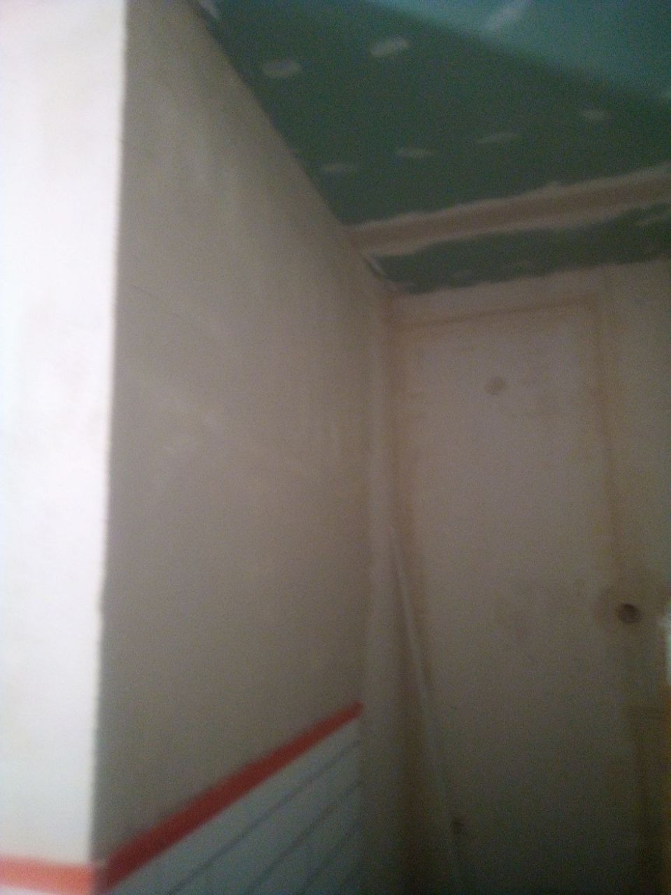 Ściana w trakcie szpachlowania, widoczne ślady po nałożonej masie, sufit z płyt gipsowo-kartonowych, fragment drzwi w tle.