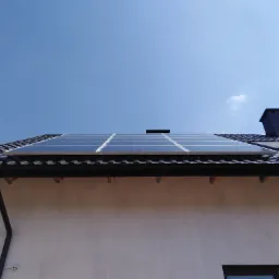 Panele słoneczne na dachu domu jednorodzinnego z ciemną dachówką i kominem na tle błękitnego nieba.