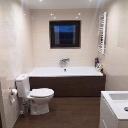 Usługi budowlano-remontowe MAR-BUD Marcin Biodrowski - Jasna łazienka z brązowymi akcentami: płytki imitujące drewno na podłodze, obudowa wanny w kolorze ciemnego drewna, beżowe płytki na ścianach z delikatnym wzorem, okno z widokiem na nocne drzewa...