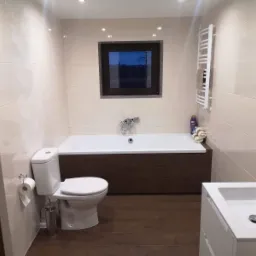 Jasna łazienka z brązowymi akcentami: płytki imitujące drewno na podłodze, obudowa wanny w kolorze ciemnego drewna, beżowe płytki na ścianach z delikatnym wzorem, okno z widokiem na nocne drzewa...