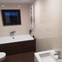 Usługi budowlano-remontowe MAR-BUD Marcin Biodrowski - Jasna łazienka z brązowymi akcentami: wanna z obudową z płytek imitujących drewno, umywalka z szafką, grzejnik drabinkowy na ścianie z beżowych płytek, okno z widokiem na nocny krajobraz.