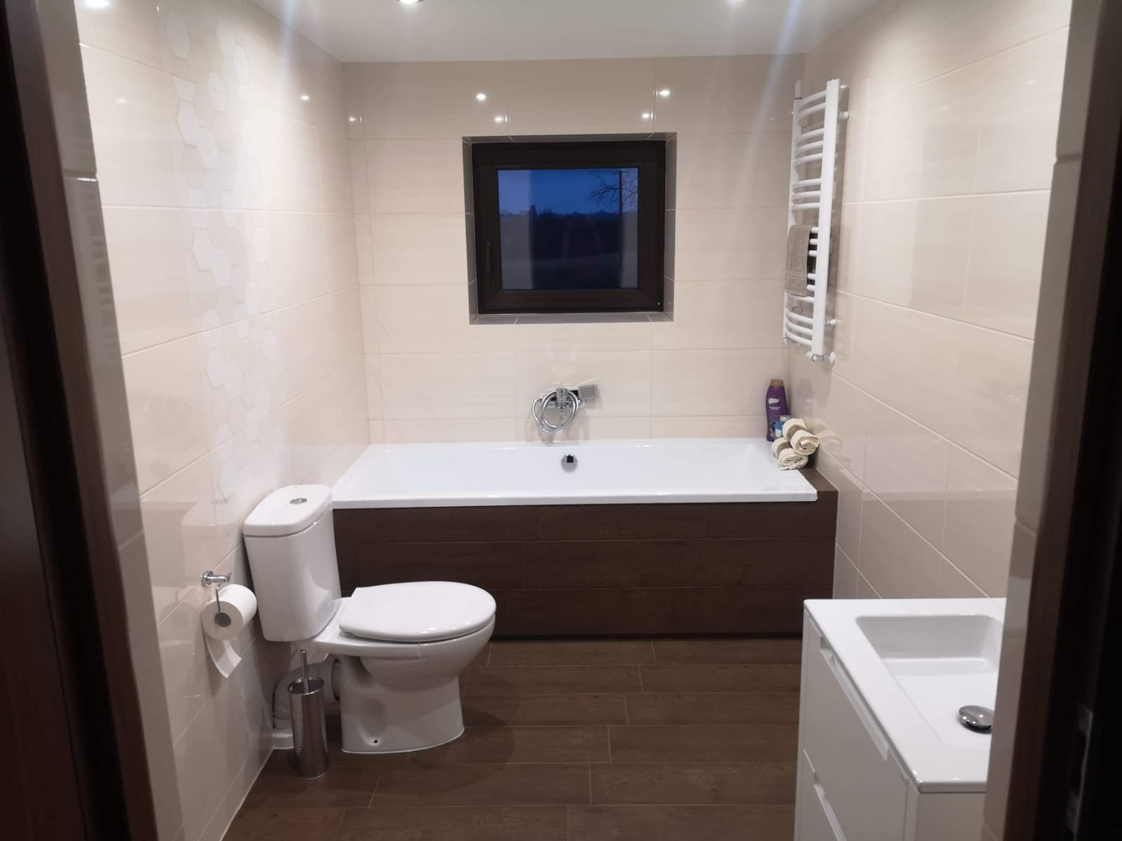 Jasna łazienka z brązowymi akcentami: płytki imitujące drewno na podłodze, obudowa wanny w kolorze ciemnego drewna, beżowe płytki na ścianach z delikatnym wzorem, okno z widokiem na nocne drzewa...