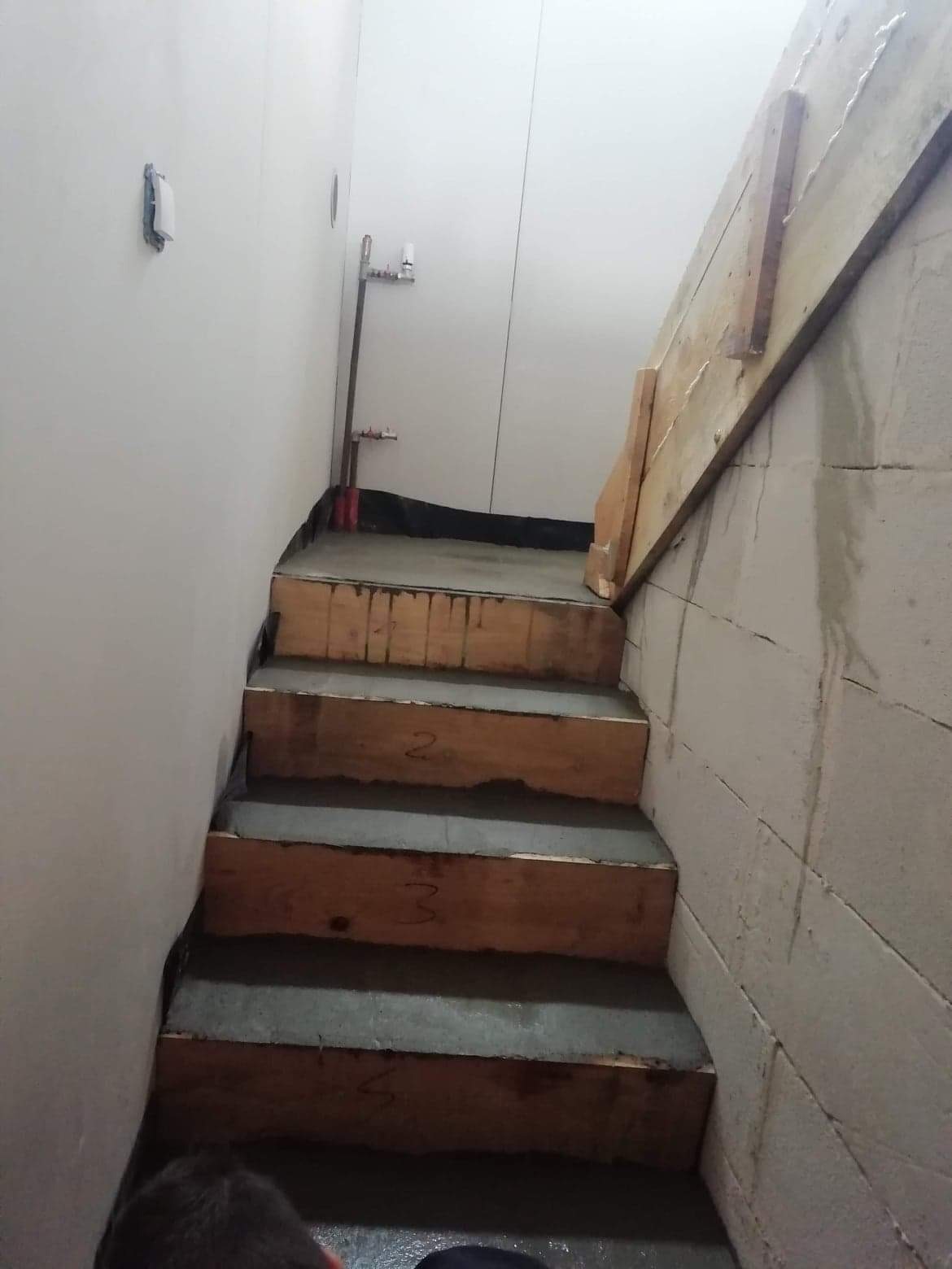 Betonowe schody w surowym stanie, z drewnianym szalunkiem i numerami na stopniach, widoczne rury instalacyjne przy ścianie.