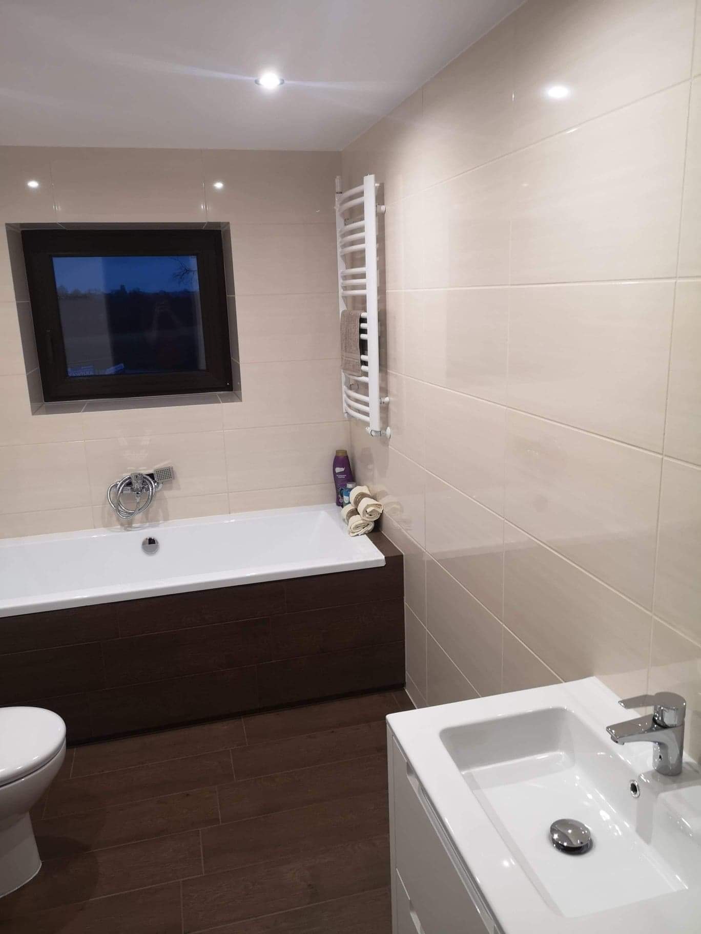 Jasna łazienka z brązowymi akcentami: wanna z obudową z płytek imitujących drewno, umywalka z szafką, grzejnik drabinkowy na ścianie z beżowych płytek, okno z widokiem na nocny krajobraz.