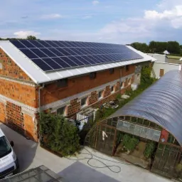 Ceglany budynek gospodarczy z panelami słonecznymi na dachu, obok szklarnie foliowe i zaparkowane busy firmy.