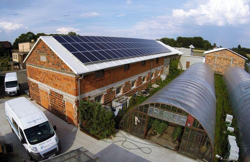 Ceglany budynek gospodarczy z panelami słonecznymi na dachu, obok szklarnie foliowe i zaparkowane busy firmy.