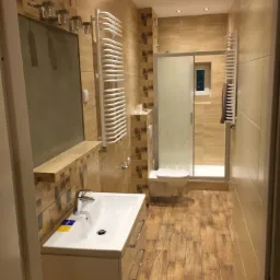 Wykończona łazienka z kabiną prysznicową, umywalką z szafką i grzejnikiem drabinkowym, wykończona płytkami imitującymi drewno na podłodze i ścianach.