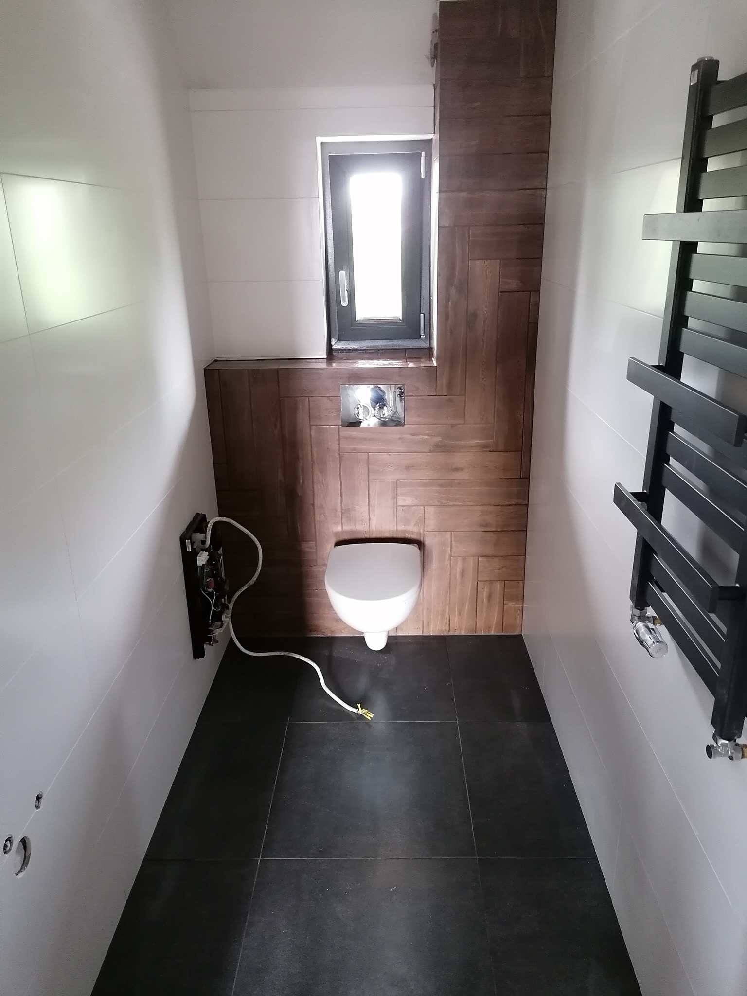 Mała łazienka w trakcie remontu: białe płytki ścienne, czarna podłoga, częściowo wykończona ściana z drewnopodobnych płytek, wisząca toaleta, grzejnik drabinkowy, widoczne przewody elektryczne.