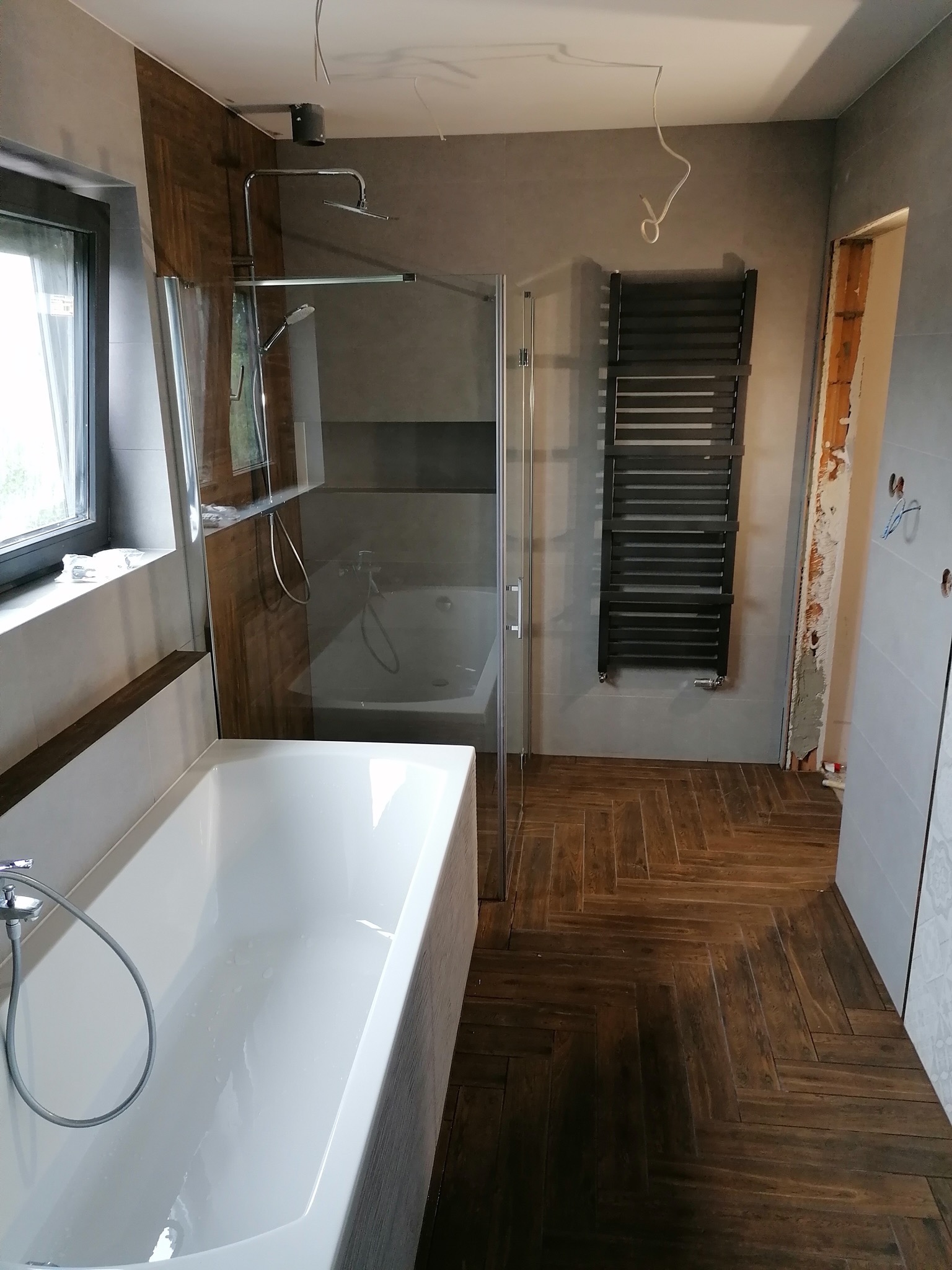Wnętrze łazienki w trakcie remontu: biała wanna, szklany parawan nawannowy, grzejnik drabinkowy na szarej ścianie, podłoga z płytek imitujących drewno ułożonych w jodełkę, widoczne kable wystające...