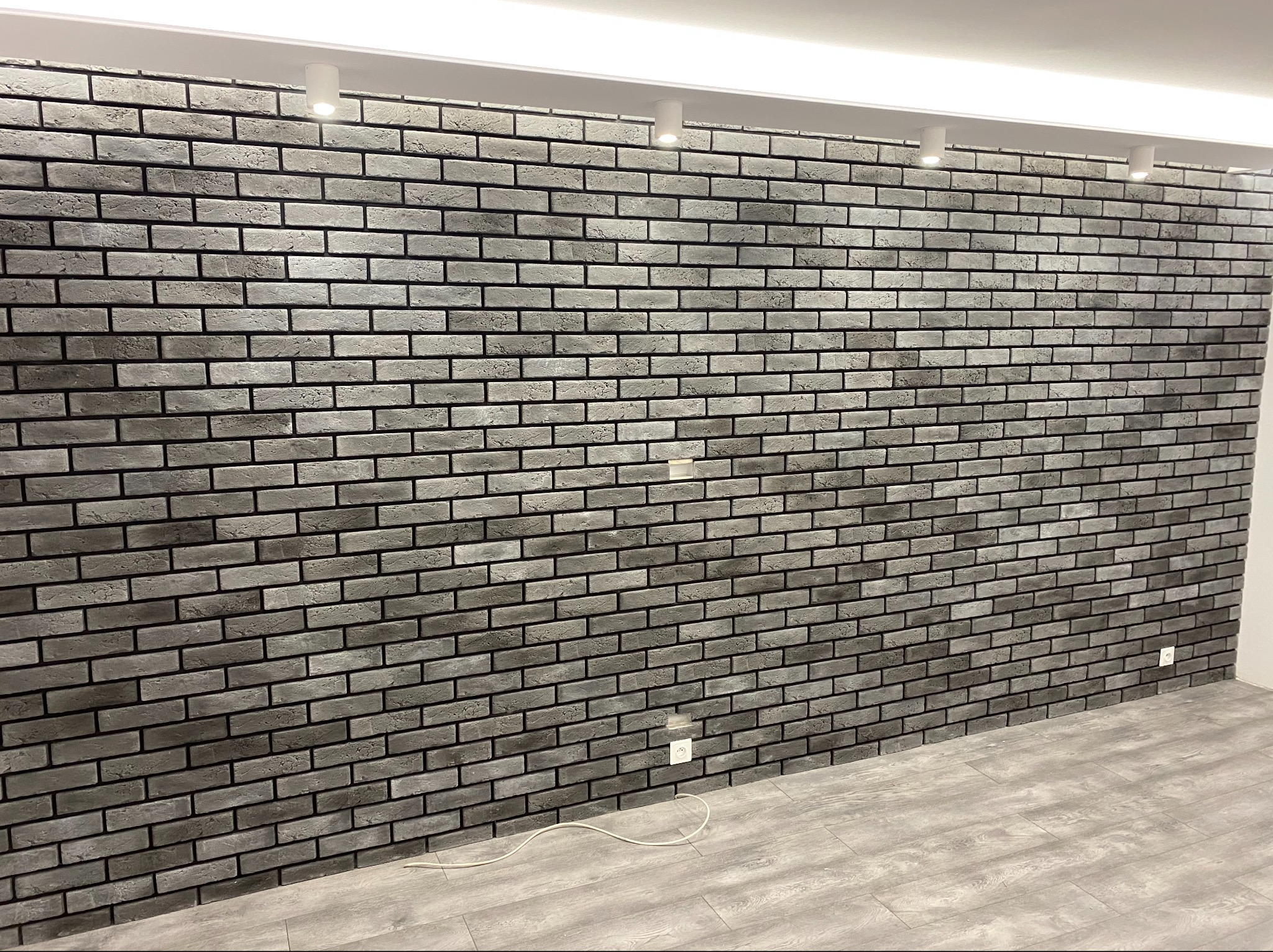 Ściana wyłożona szarymi cegłami klinkierowymi z czarną fugą, oświetlona punktowymi lampami sufitowymi, widoczny fragment podłogi z paneli imitujących drewno i gniazdko elektryczne.