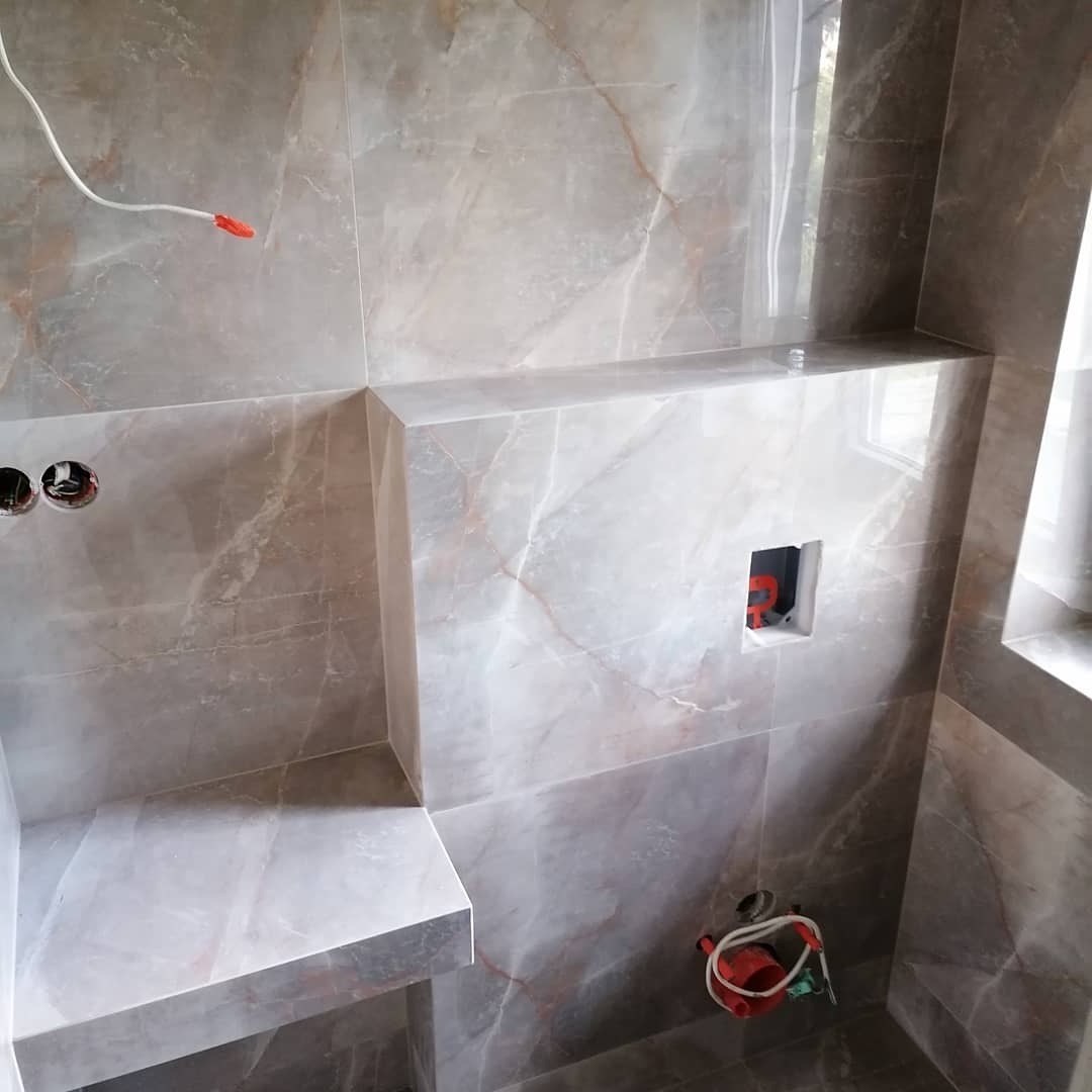 Wnętrze łazienki w trakcie wykańczania: marmurowe płytki ścienne z widocznymi otworami na gniazdka elektryczne i instalację wodną, niedokończona półka i wystający kabel elektryczny.