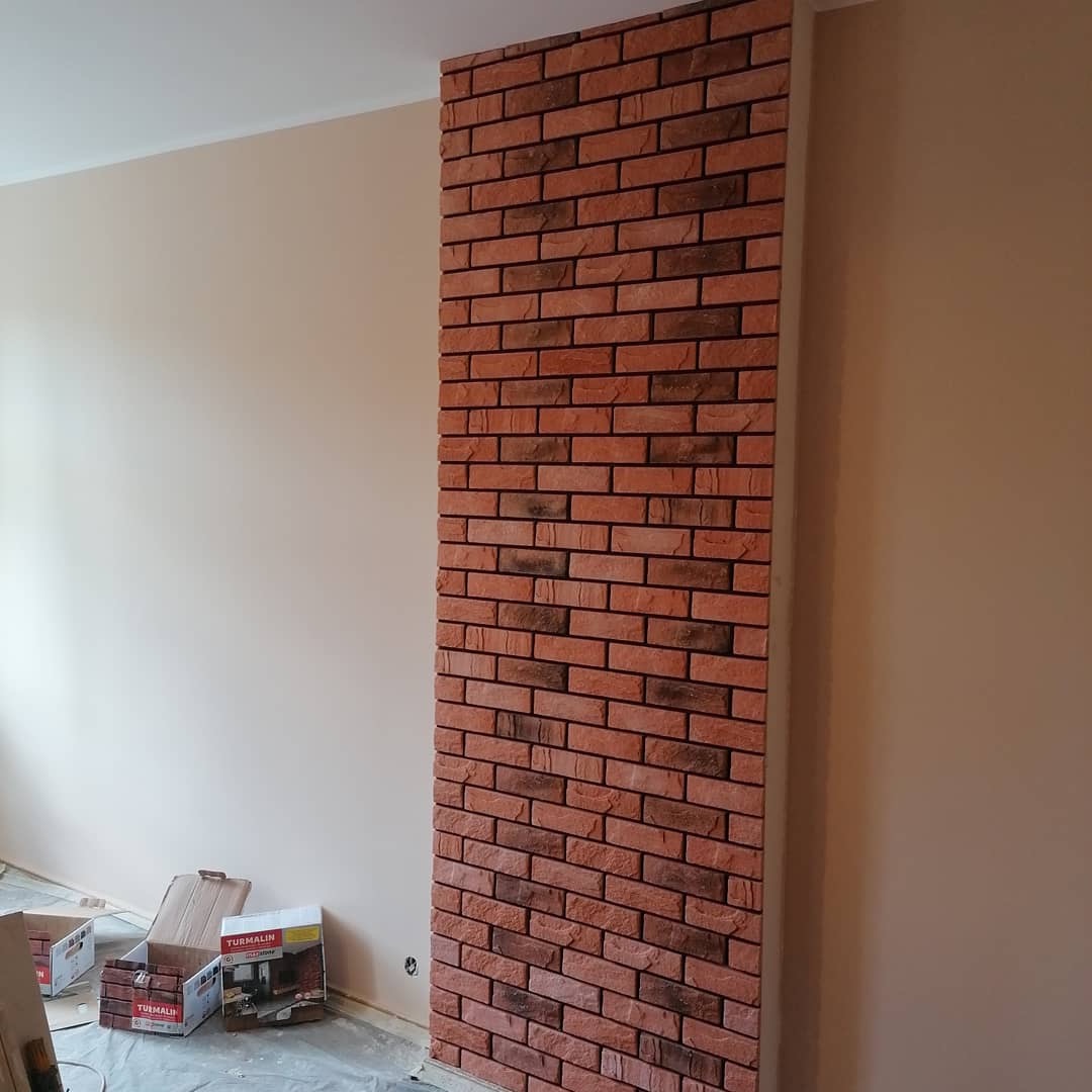 Fragment ściany z dekoracyjną okładziną z czerwonej cegły, widoczne kartony z okładziną na podłodze, surowe wykończenie ścian w tle.