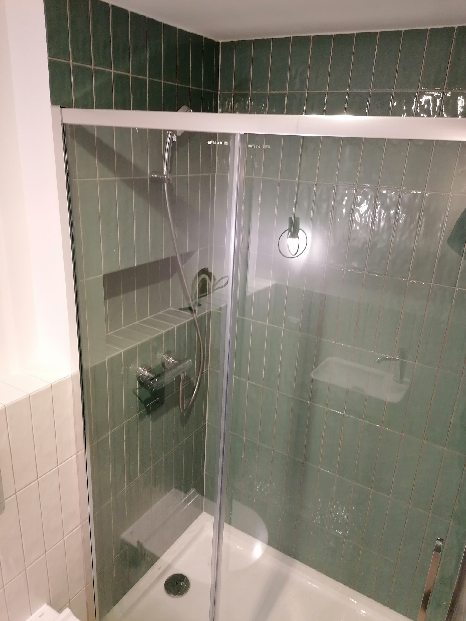 Kabina prysznicowa z zielonymi, pionowo ułożonymi płytkami, widoczny zestaw prysznicowy i wnęka w ścianie wyłożona płytkami.