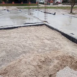 Wylany fundament domu z wystającymi prętami zbrojeniowymi, częściowo zasypany piaskiem, widoczne czarne folie izolacyjne.