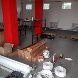 Wnętrze remontowanego pomieszczenia z czerwonymi filarami, widoczne kable elektryczne, narzędzia, kartony z elementami wykończeniowymi, drabina i grzejniki żeliwne pod oknami.