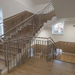 Klatka schodowa w budynku z drewnianymi schodami i metalowymi balustradami, widok z dołu na górę, okna z widokiem na drzewa na zewnątrz.