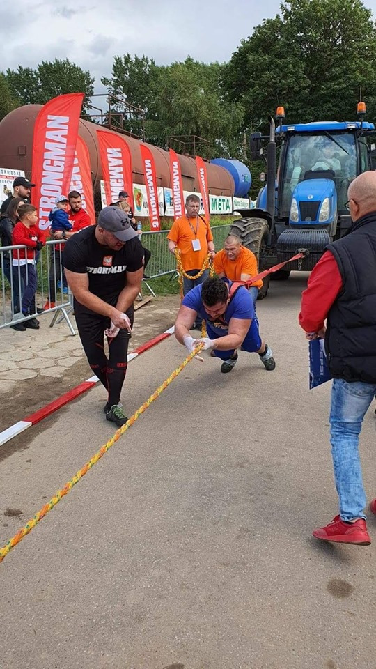 Zawody strongman: mężczyzna w niebieskiej koszulce ciągnie linę, opierając się na rękach, z pomocą asekurujących osób w pomarańczowych koszulkach, w tle wagon kolejowy i traktor.