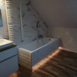 Nowoczesna łazienka na poddaszu z marmurowymi płytkami, wanną obłożoną białymi kafelkami typu metro i drewnopodobną podłogą, oświetlona listwą LED pod wanną.