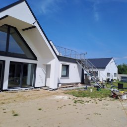 Termo-Dom Zbigniew Józefowicz - Nowoczesny dom z białymi ścianami i czarnymi ramami okiennymi, widoczne elementy rusztowania i przygotowania do dalszych prac wykończeniowych elewacji, błękitne niebo w tle.