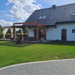 Nowoczesny dom jednorodzinny z drewnianą pergolą i białymi zasłonami na tarasie, zadbany trawnik, brukowany podjazd, błękitne niebo z obłokami.