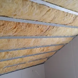 Skośny sufit poddasza z izolacją z wełny mineralnej zamontowaną między metalowymi profilami, częściowo zakryty płytami kartonowo-gipsowymi, widoczne gniazdka elektryczne.