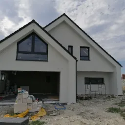 Nowoczesny dom jednorodzinny z białą elewacją w trakcie malowania, widoczne rusztowanie i materiały budowlane, ciemna dachówka i okna.