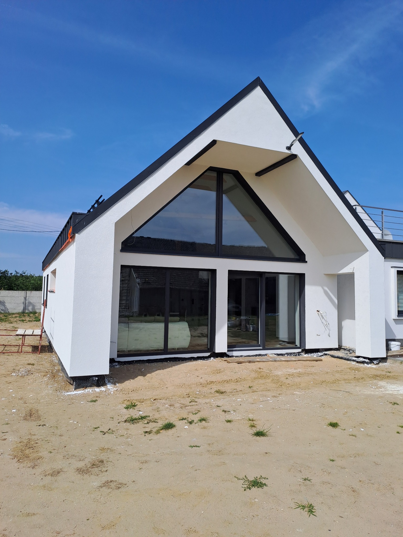 Nowoczesny dom jednorodzinny z białą elewacją i dużymi przeszkleniami, widok z zewnątrz w słoneczny dzień, teren wokół domu w trakcie zagospodarowywania.