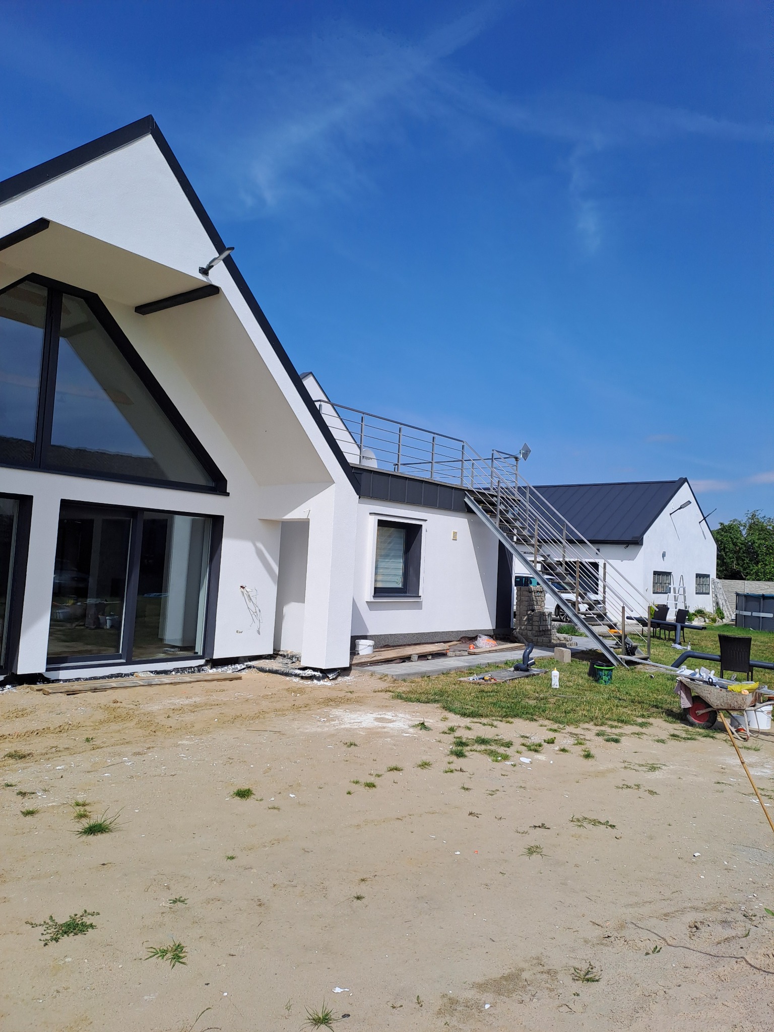 Nowoczesny dom z białymi ścianami i czarnymi ramami okiennymi, widoczne elementy rusztowania i przygotowania do dalszych prac wykończeniowych elewacji, błękitne niebo w tle.