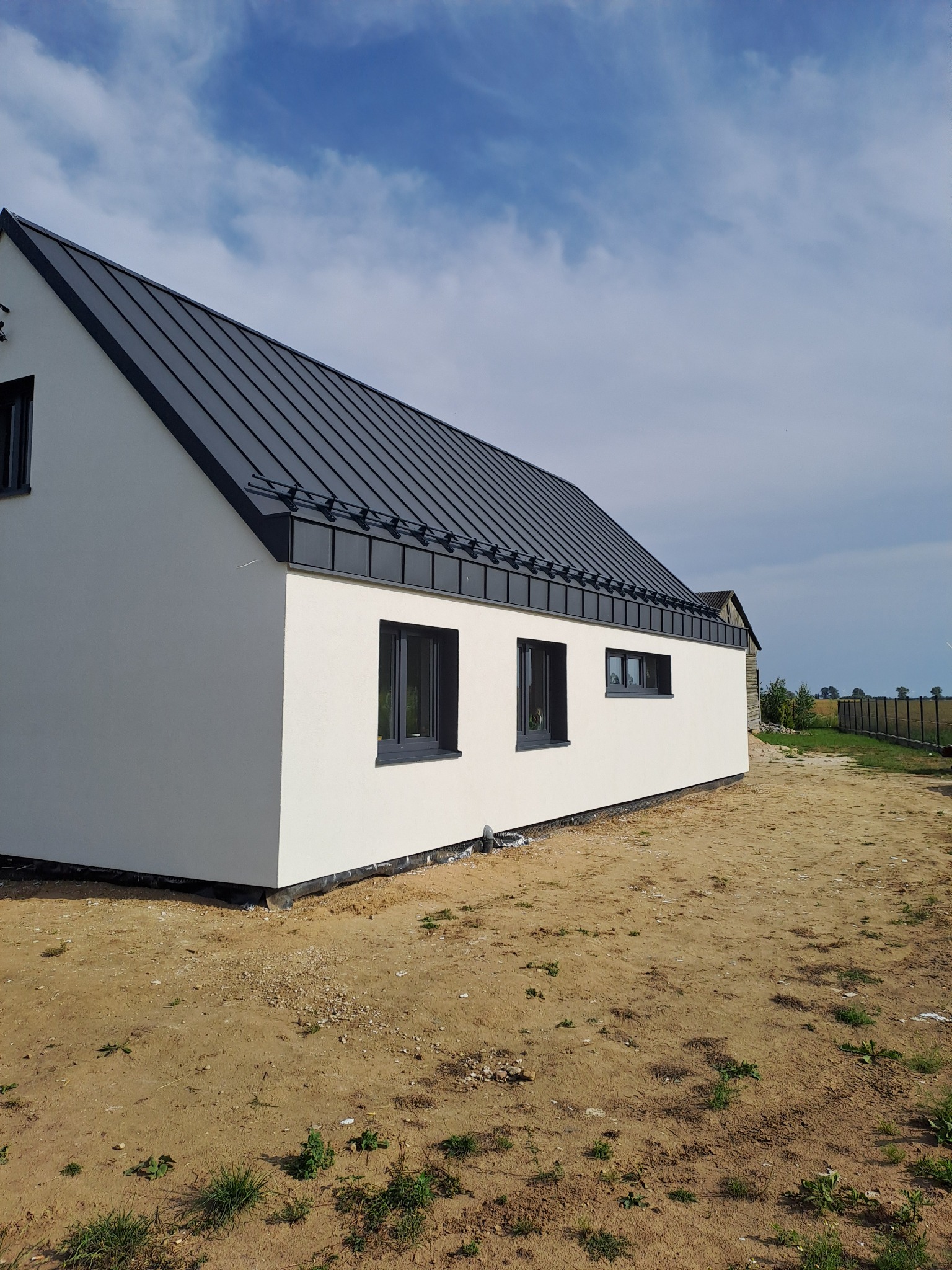 Nowoczesny, biały dom jednorodzinny z grafitowym dachem z blachy na rąbek stojący i ciemnymi oknami, na działce z widoczną ziemią i fragmentami trawy, w tle zabudowania gospodarcze i błękitne niebo.