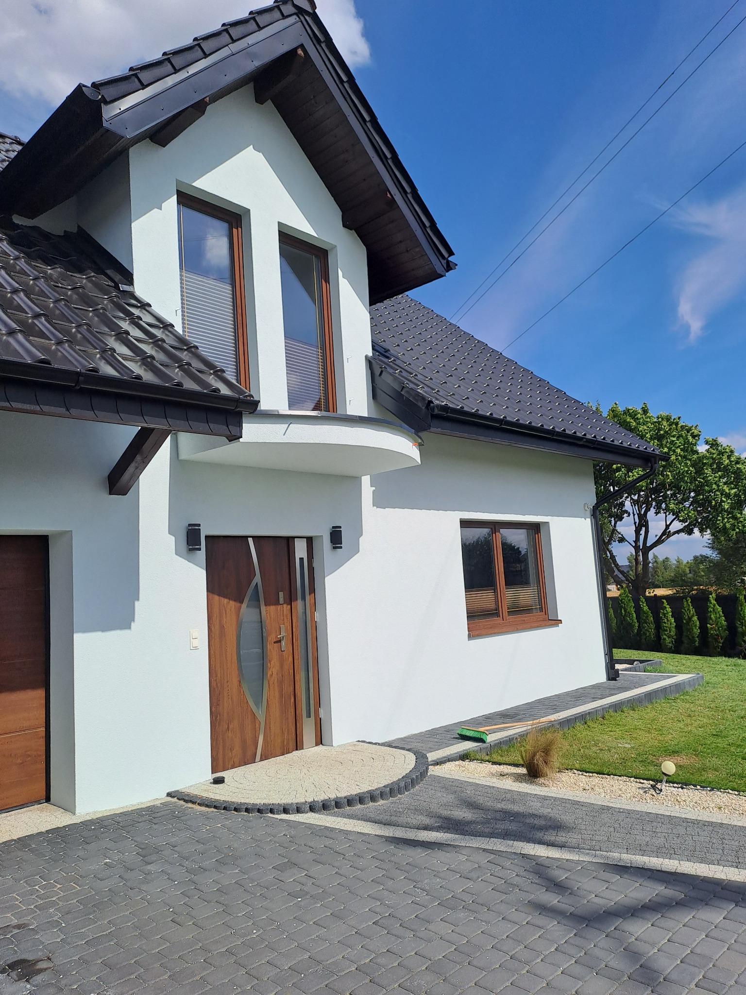 Nowoczesny dom jednorodzinny z jasną elewacją, ciemną dachówką i drewnianymi elementami wykończeniowymi, widok z zewnątrz na elewację frontową i podjazd z kostki brukowej.