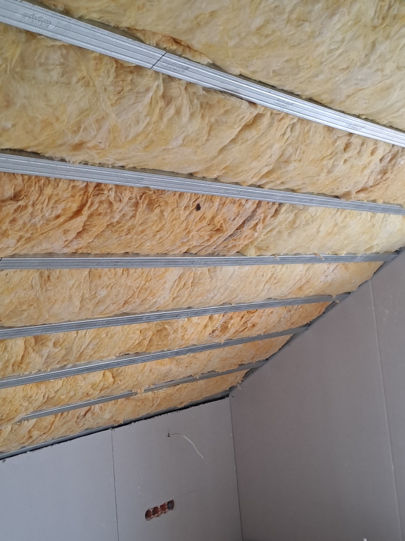 Skośny sufit poddasza z izolacją z wełny mineralnej zamontowaną między metalowymi profilami, częściowo zakryty płytami kartonowo-gipsowymi, widoczne gniazdka elektryczne.