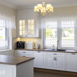 Stolar Group Rafał Kryger - Jasna kuchnia z białymi frontami, drewnianym blatem, złotą armaturą i koronkowymi firankami w oknach, widok na zieleń za oknem.