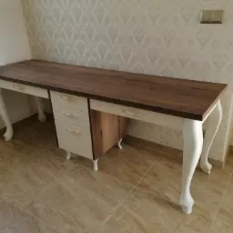 Biurko z drewnianym blatem i białymi, giętymi nogami w stylu retro, z szufladami i szafką, na tle ściany z wzorzystą tapetą i podłogi z płytek.