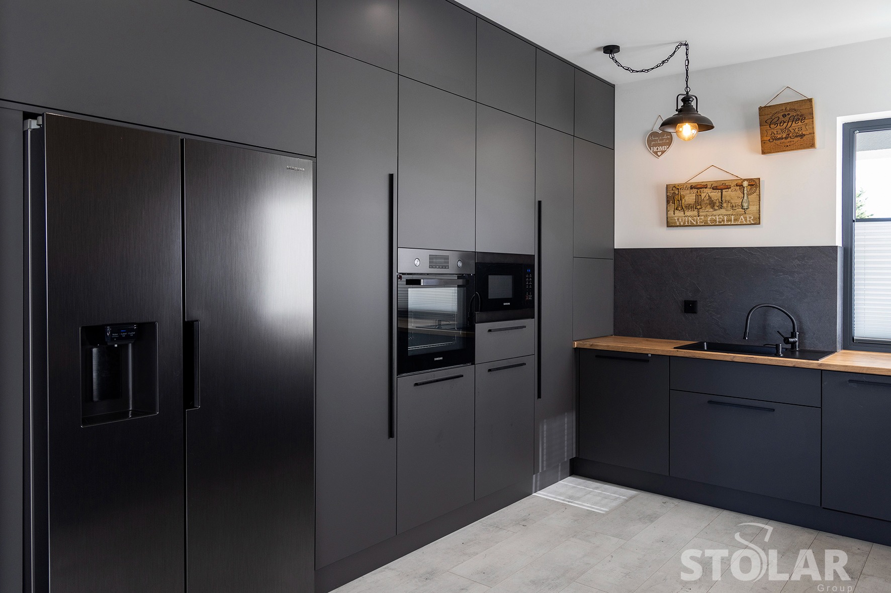 Nowoczesna kuchnia w odcieniach szarości z zabudową sięgającą sufitu, sprzęt AGD marki Samsung wkomponowany w zabudowę, drewniany blat i industrialna lampa wisząca.