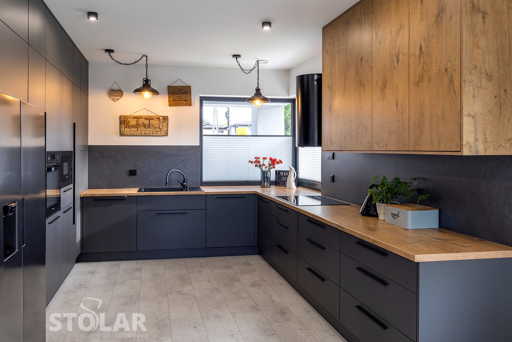 Nowoczesna kuchnia w odcieniach szarości i drewna z wiszącymi lampami w stylu industrialnym, drewnianym blatem i dekoracjami ściennymi.