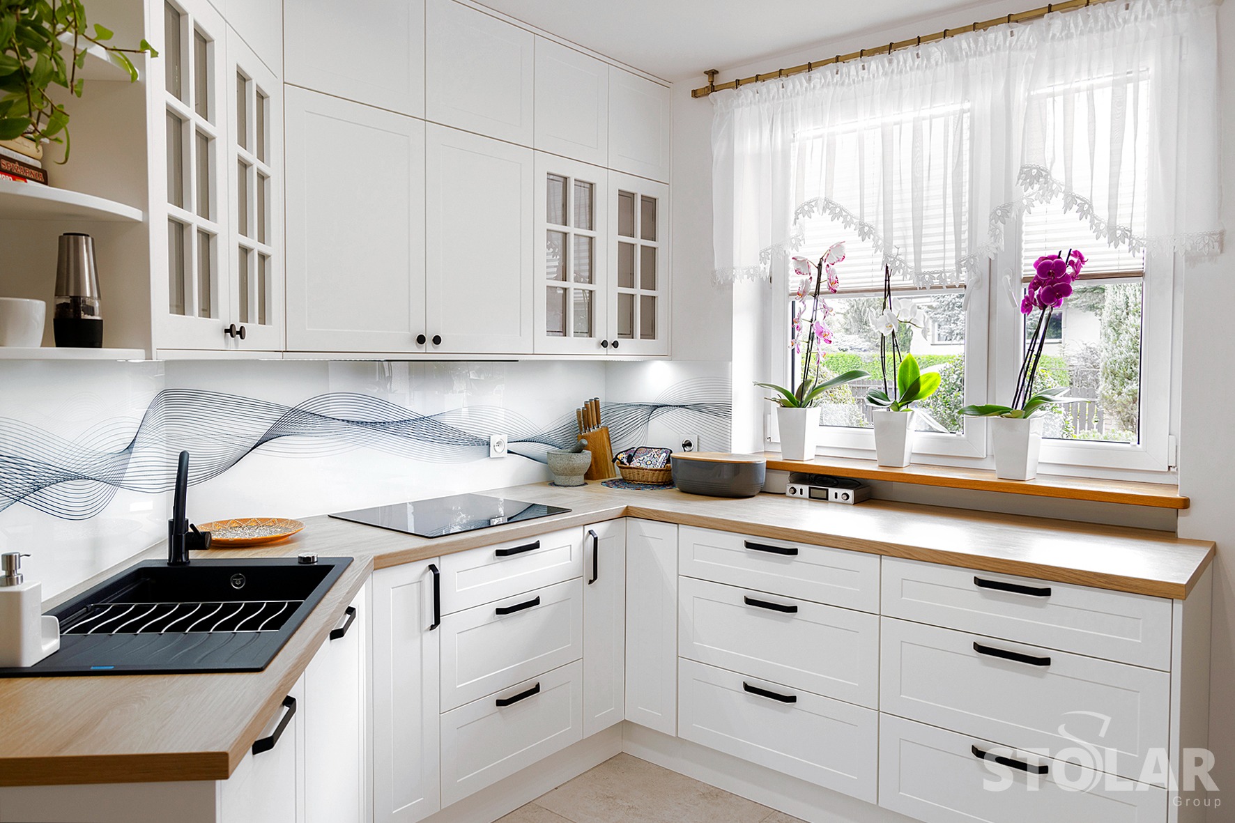 Jasna kuchnia z białymi frontami szafek, drewnianym blatem i czarnym zlewozmywakiem, ozdobiona falistym wzorem na szklanym panelu nad blatem oraz trzema storczykami w doniczkach na parapecie okna.