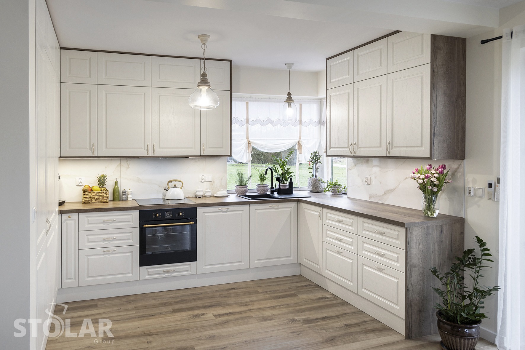 Jasna, klasyczna kuchnia z drewnianymi blatami i białymi frontami, oświetlona wiszącymi lampami, z widokiem na ogród za oknem.