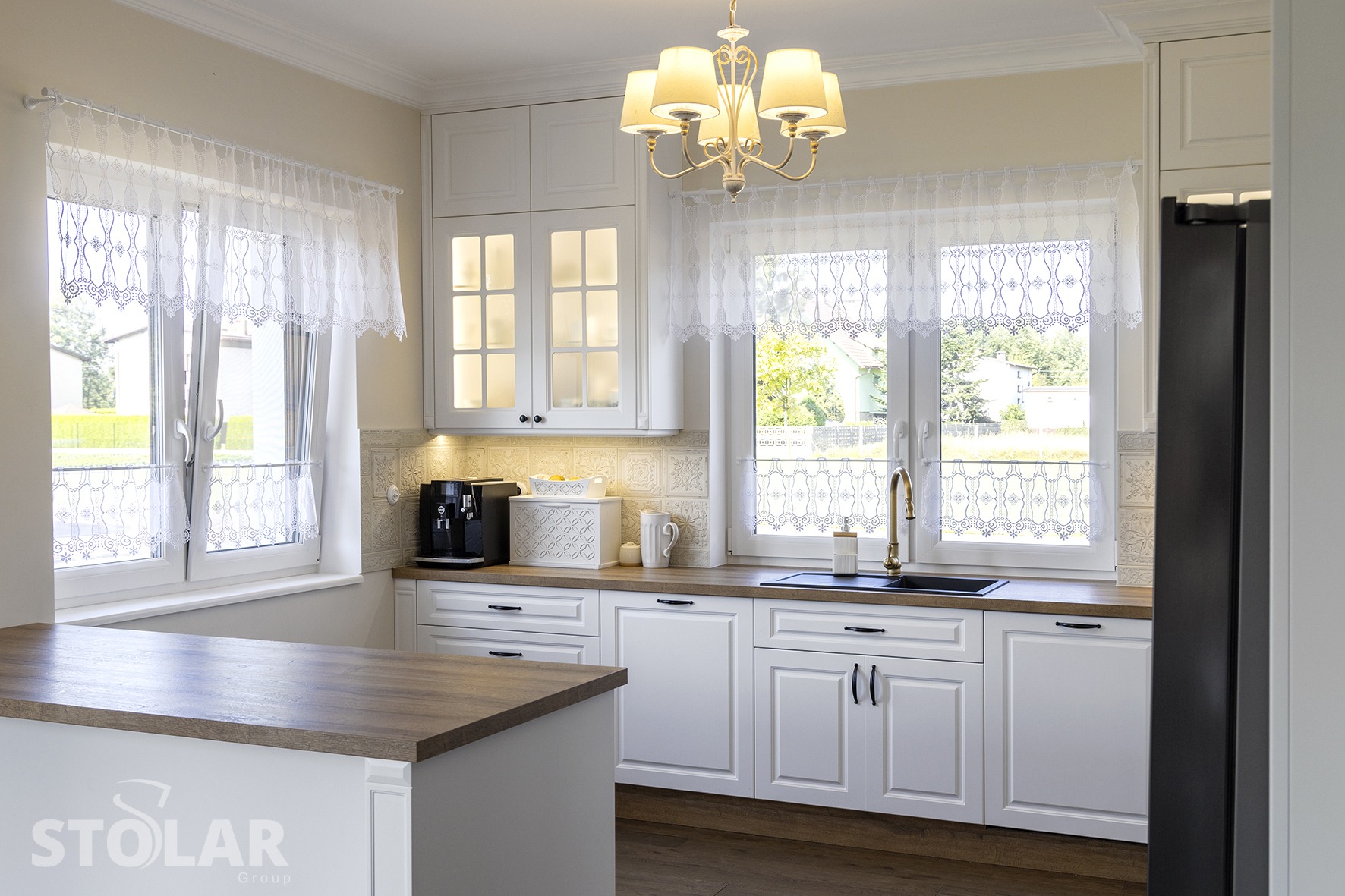 Jasna kuchnia z białymi frontami, drewnianym blatem, złotą armaturą i koronkowymi firankami w oknach, widok na zieleń za oknem.