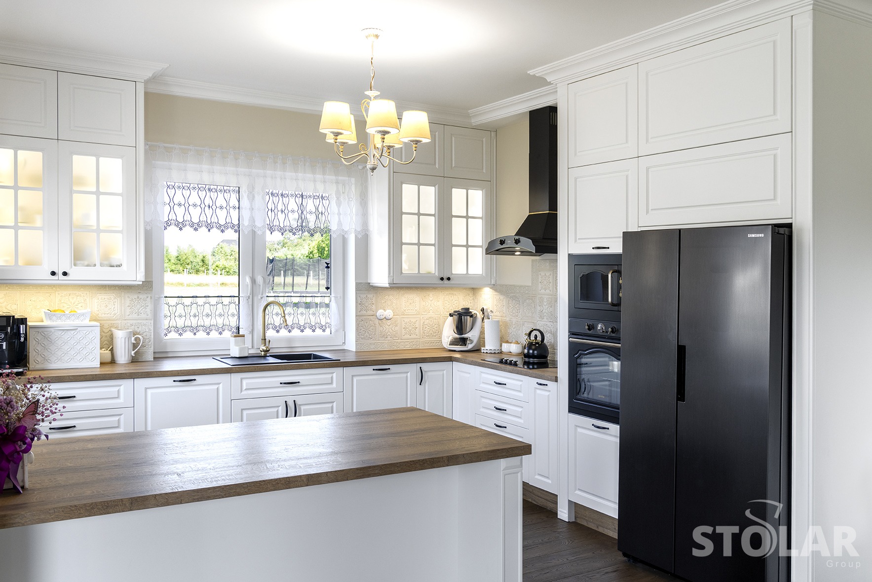 Klasyczna kuchnia w bieli z drewnianym blatem wyspy, czarnym sprzętem AGD i dekoracyjną firanką w oknie.