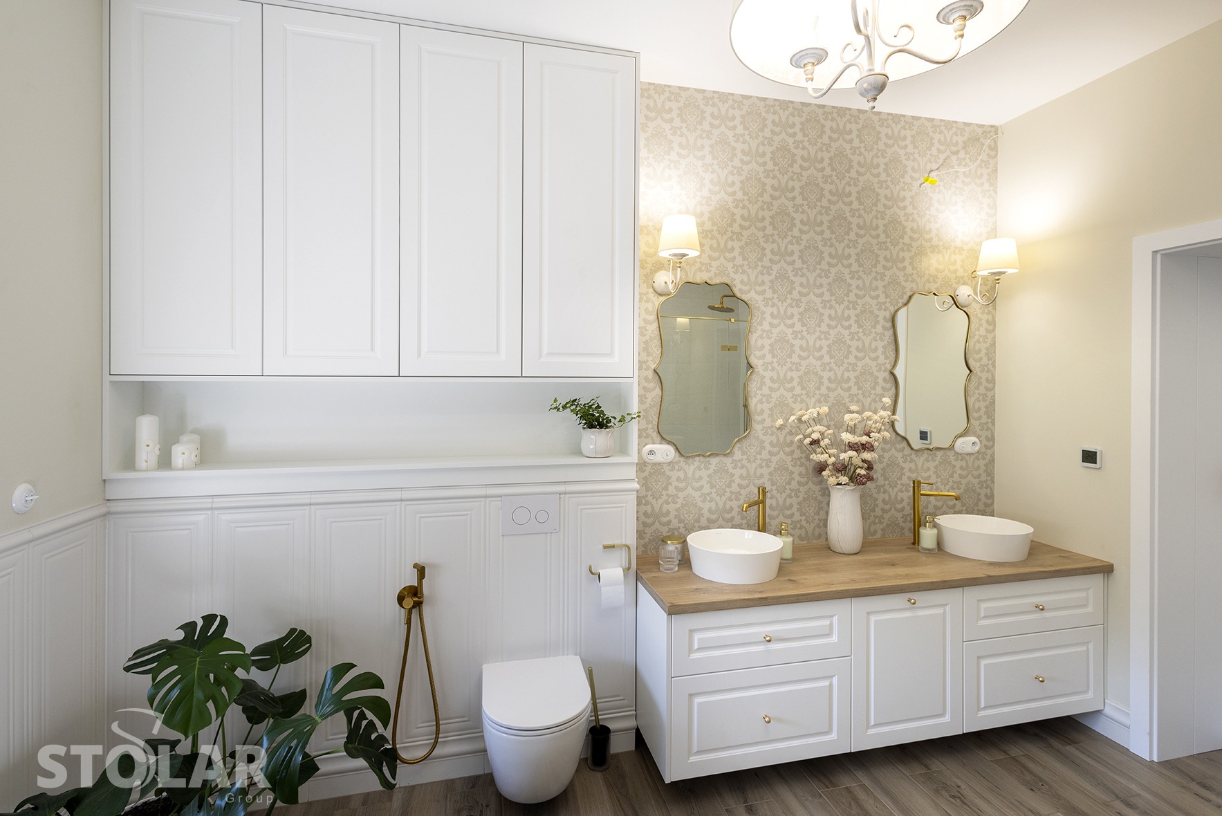Jasna łazienka z białymi szafkami pod umywalkami, drewnianym blatem, dekoracyjną tapetą, złotą armaturą i wiszącymi szafkami na ścianie.