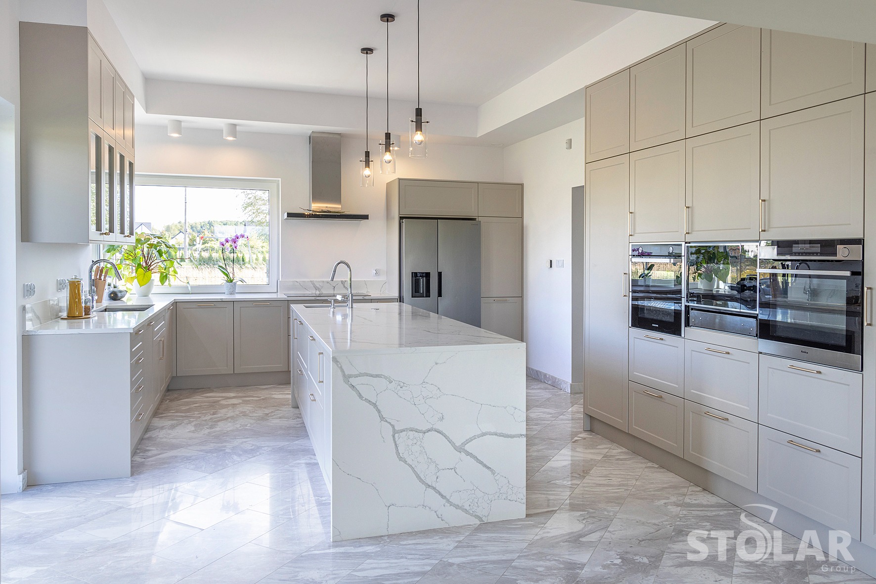Jasna kuchnia z wyspą i marmurowym blatem, widok na okno z roślinami, nowoczesne oświetlenie wiszące nad wyspą, zabudowa kuchenna z piekarnikami.