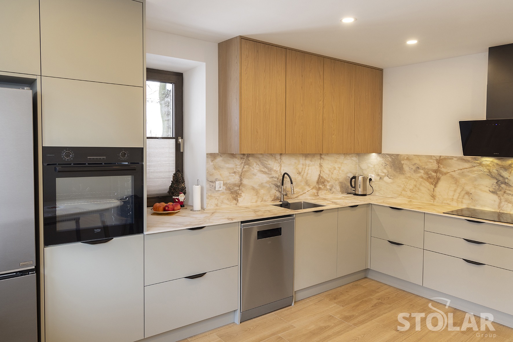 Nowoczesna kuchnia z beżowymi szafkami, drewnianymi górnymi szafkami, marmurowym blatem i backsplashiem, srebrnymi urządzeniami i drewnianą podłogą. W oknie widać dekorację świąteczną.